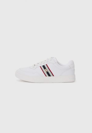 Zapatilla blanca con parte superior texturizada, con rayas horizontales en negro y rojo, diseño de cordones y suela de goma plana. Detalle del logo en dorado.