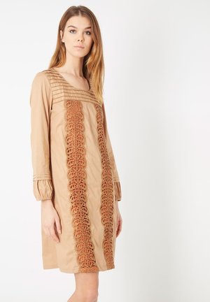 Vestido beige de mangas largas, con un trenzado texturizado en los hombros y un intrincado detalle de encaje en la parte delantera. Textura de tela suave.