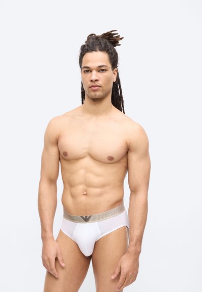 Μυώδης άνδρας με μακριά dreadlocks που στέκεται μπροστά σε λευκό φόντο, φορώντας λευκά εσώρουχα με μπεζ ριγωτή μέση.