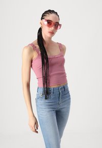 Różowy prążkowany crop top z koronkowym wykończeniem, w połączeniu z jasnoniebieskimi jeansami z wysokim stanem. Modelka nosi przezroczyste różowe okulary przeciwsłoneczne.