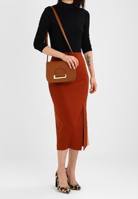 Brun læder crossover taske med detaljer i guldtonet hardware. Modellen bærer en sort top, en rustfarvet midi-nederdel med en sideslids og højhælede sko med leopardprint.