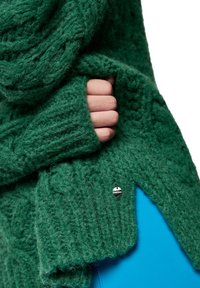 Grüner gestrickter Schal mit einem strukturierten Muster und einem silbernen Akzentknopf. Gefaltet mit einer Hand, die den Stoff hält, vor einem blauen Hintergrund.