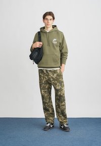 Jauns cilvēks ar olīvu zaļu Nike kapuci, kamuflāžas biksēm, melniem apaviem un melnu plecu somu pret vienkāršu fonu.