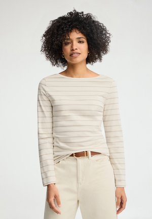 Femme aux cheveux bouclés portant une chemise beige à manches longues rayée rentrée dans un pantalon crème avec une ceinture marron, debout devant un fond uni.