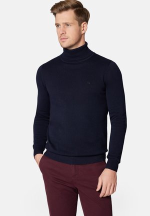 Ciemnoniebieski sweter typu golf w dopasowanym fasonie, z dzianinową teksturą, z prążkowanymi mankietami i dołem. Połączony z bordowymi spodniami. Widoczny mały detal logo.
