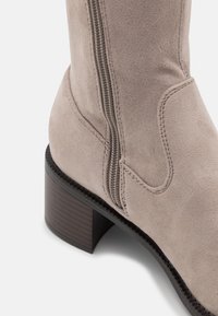 Botín de ante beige hasta la rodilla con cremallera lateral, punta redondeada y un tacón de bloque de madera texturizado. Detalle de costura cerca de la cremallera.