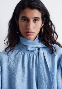 Himmelblaue, zerknitterte Bluse mit hohem Kragen und schalähnlichem Detail. Mit voluminösen Ärmeln und subtiler Textur. Goldene Creolen.