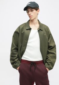 Olīvu zaļš bomber tipa mētelis ar elastīgām aprocēm, baltā topa bez piedurknēm un bordo sporta biksēm ar auklu. Nike logo uz mēteļa un biksēm.