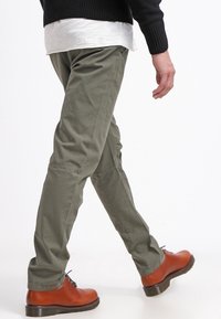 Person som går iförd olivgröna byxor, svart tröja, vit skjorta och bruna läderskor på en vit bakgrund.