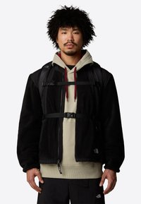 Casaco de fleece preto com alças ajustáveis, sobre um hoodie bege. Apresenta fechos de correr e logótipos. O modelo também tem uma mochila.