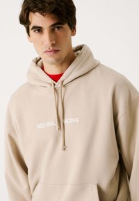 Sudadera beige hecha de tela suave, con un bolsillo frontal y capucha con cordón. Muestra "RED BULL RACING" en texto blanco a lo largo del pecho.