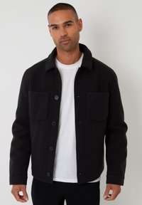 Sort uldblandingsjakke med knaplukning foran, to brystlommer og krave, iført over en hvid t-shirt, mod en ensfarvet baggrund.