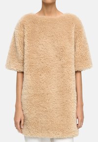 Osoba w beżowym, krótkim swetrze o puszystej fakturze, który sięga poza biodra, w połączeniu z białymi spodniami, stoi na tle prostego tła.