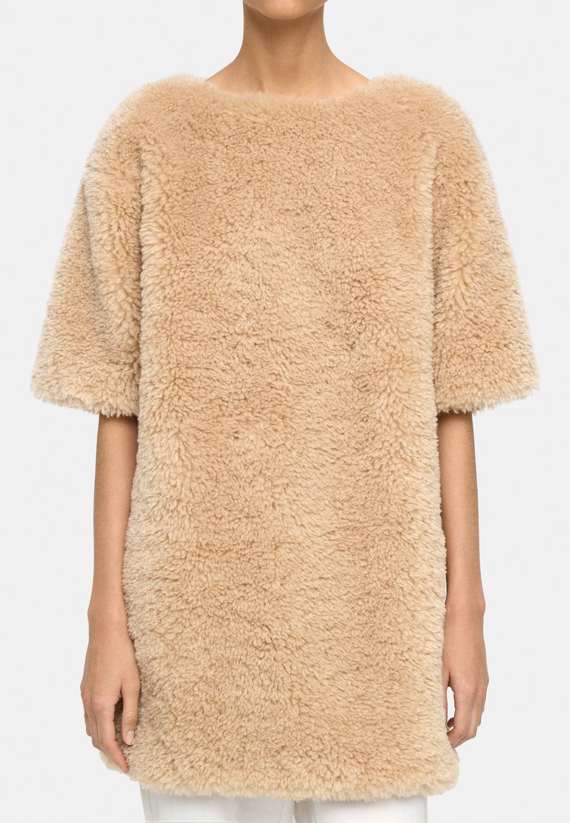 Osoba w beżowym, krótkim swetrze o puszystej fakturze, który sięga poza biodra, w połączeniu z białymi spodniami, stoi na tle prostego tła.