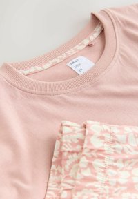 Haut en coton rose doux avec un col arrondi et un tissu à motif floral assorti plié dessus, montrant une étiquette blanche à l'intérieur du col.