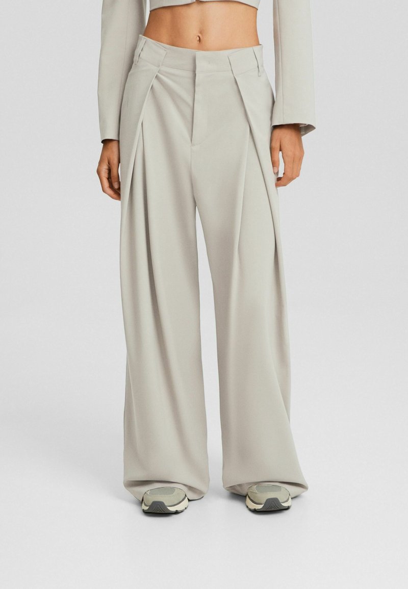 Pantaloni ampi grigi chiari con pences frontali, vita sartoriale e una texture di tessuto liscia. Indossati con scarpe da ginnastica di colore neutro.