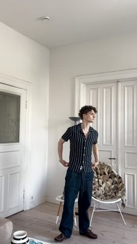 Camisa de manga curta às riscas em azul marinho e branco, calças largas azul-escuras, de pé numa sala iluminada com chão de madeira clara e uma cadeira decorativa.