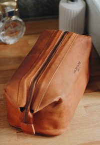 Brown leather toiletry bag with a rectangular shape, zipper closure, and subtle stitching details. Embossed text on one side.
