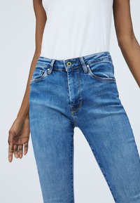 Jeans blu slim fit indossati da una persona con pelle di media tonalità marrone e top bianco senza maniche, visibili dalla vita a metà coscia su uno sfondo semplice.