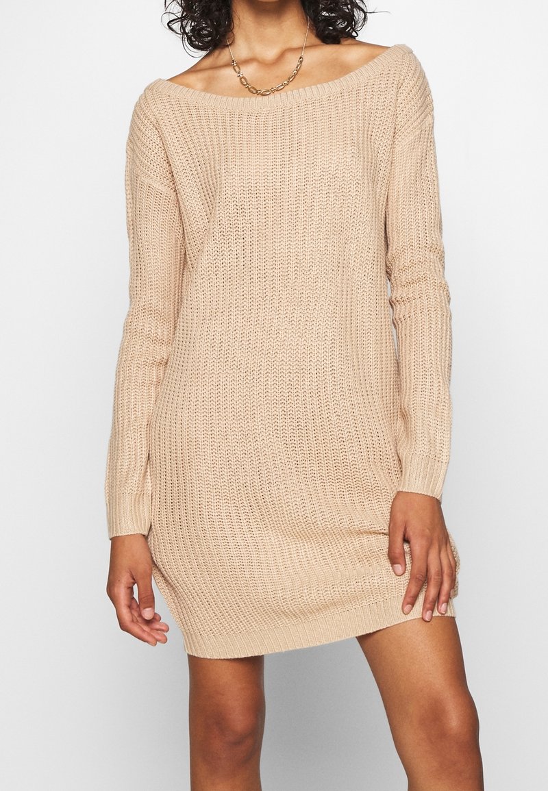 Femme portant une robe-pull beige en maille épaules dénudées avec manches longues et un collier chaîne doré sur fond blanc.