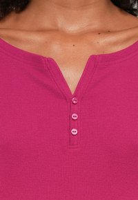 Fuchsia ribbet top med V-udskæring, der har tre pink knapper ved placket. Glat tekstur og stof af medium vægt.