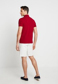Röd polo-shirt med korta ärmar, smal passform. Matchad med vita shorts och mörka skor. Bakgrunden är helt vit.