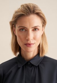 Primo piano di una persona con capelli biondi fino alle spalle che indossa una camicia blu scuro con colletto, chiusura con bottoni e tessuto dalla texture liscia.