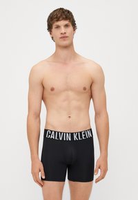 Boxer da uomo neri con una morbida consistenza e una vita elasticizzata con la scritta in bianco "CALVIN KLEIN". Modello posato con il corpo superiore nudo.