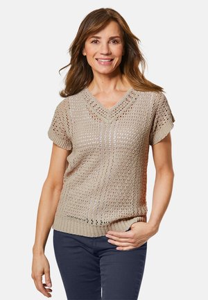 Beiger V-Ausschnitt-Strickpullover mit kurzen Ärmeln, der eine strukturierte, offene Maschenware und einen gerippten Saum aufweist. Kombiniert mit navyblauen Hosen.