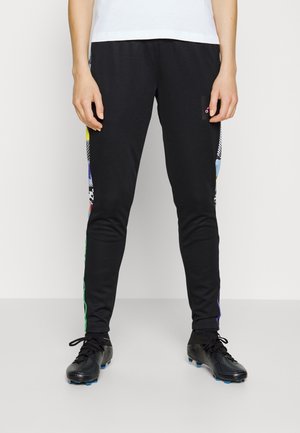Personne portant un pantalon de sport noir avec des panneaux latéraux colorés et des crampons noirs, debout devant un fond clair uni.