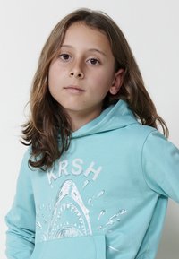 Tirkīza kapuce ar priekšējo kabatu, kas rotāta ar baltu haizivju grafiku un "KRSH" tekstu. Gluda auduma, ikdienišķs piegriezums un regulējama kapuce.