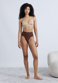 Μπεζ σουτιέν και καφέ ψηλόμεσο εσώρουχο από λείο, ελαστικό ύφασμα. Το μοντέλο στέκεται σε ανοιχτό χαλί με απαλή υφή.