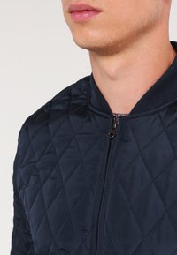 Chaqueta bomber acolchada color navy con cierre frontal, cuello acanalado y patrón de diamante. Textura suave con un detalle de tela interior en contraste.