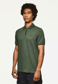 Mężczyzna z kręconymi włosami, ubrany w oliwkową koszul�ę polo z krótkim rękawem, wykończoną czarnymi akcentami i małym logo na piersi, stojący na jednolitym tle.