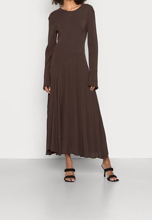 Femme portant une robe longue à manches longues, de couleur marron foncé, avec une jupe fluide et des sandales noires à talons hauts avec des bretelles à volants.