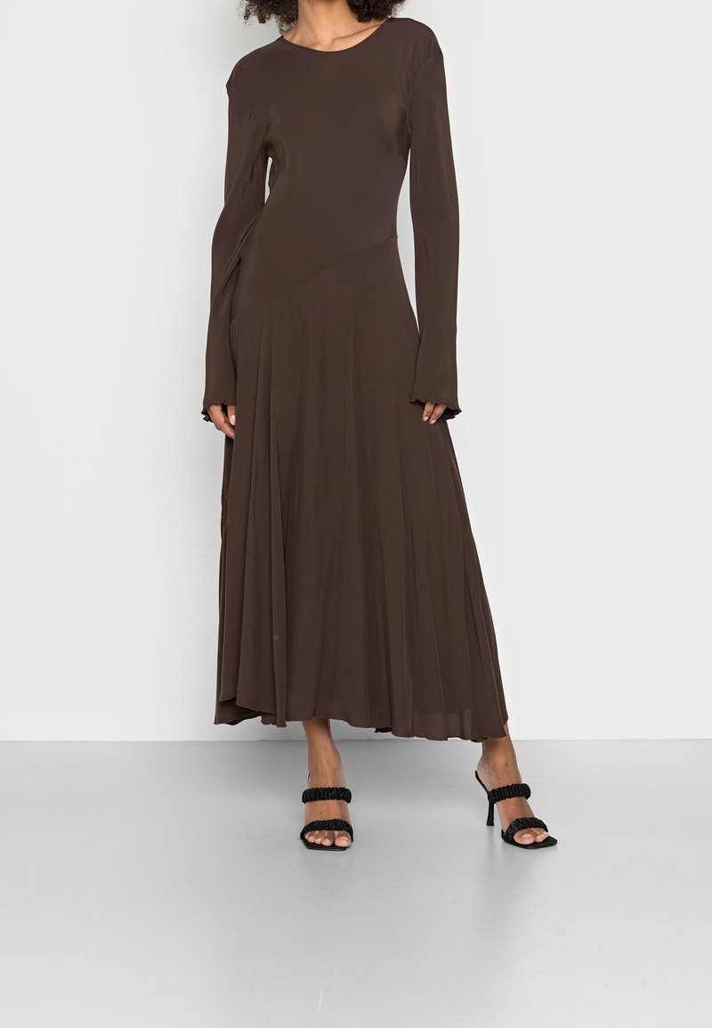 Femme portant une robe longue à manches longues, de couleur marron foncé, avec une jupe fluide et des sandales noires à talons hauts avec des bretelles à volants.