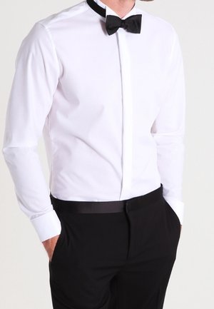 Hombre con camisa blanca de vestir, corbatín negro y pantalones negros, con las manos en los bolsillos, de pie frente a un fondo liso.