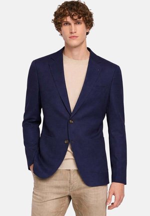 Giovane uomo con capelli ricci che indossa un blazer blu navy sopra un maglione beige e pantaloni marrone chiaro su uno sfondo bianco uniforme.