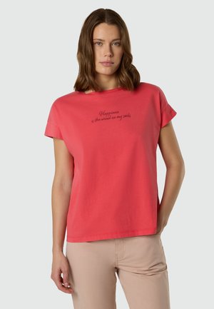 Donna con capelli castani fino alle spalle che indossa una maglietta rosa corallo con la scritta "La felicità è il vento nelle mie vele" e pantaloni beige.