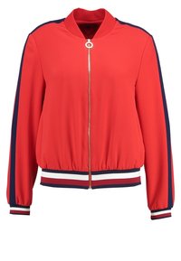 Röd bomberjacka med dragkedja framtill, marinblå sidskenor och ribbade muddar och fåll. Tillverkad av slät, lättviktsväv.