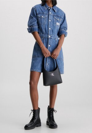 Femme portant une robe en jean retroussée, tenant un sac à main noir Calvin Klein, et des bottes de combat noires à lacets, debout contre un fond uni.