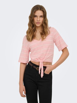 Jeune femme aux cheveux ondulés porte un crop top rose rayé à manches courtes noué et un pantalon noir avec une ceinture tressée, debout devant un fond uni.