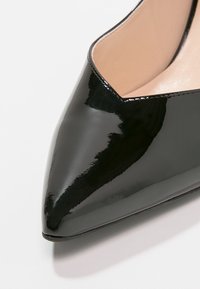 Gros plan sur une chaussure à bout pointu en cuir verni noir brillant avec doublure intérieure beige sur fond blanc.