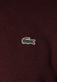 Burgundypolo-skjorta i tyg med ribbad struktur, med en broderad grön Krokodil-logotyp i vitt runtom.