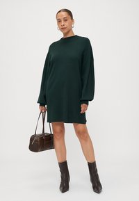 Hosszú ujjú, sötétzöld kötött ruha magas nyakkivágással. Kényelmes szabású és puha textúrájú, sötétbarna táskával és térd fölé érő csizmával párosítva.
