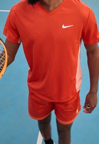 Sada výkonného sportovního oblečení v jasně oranžové barvě, zahrnující tričko s krátkým rukávem ve v-neck stylu s bílými detaily a odpovídající texturované šortky.