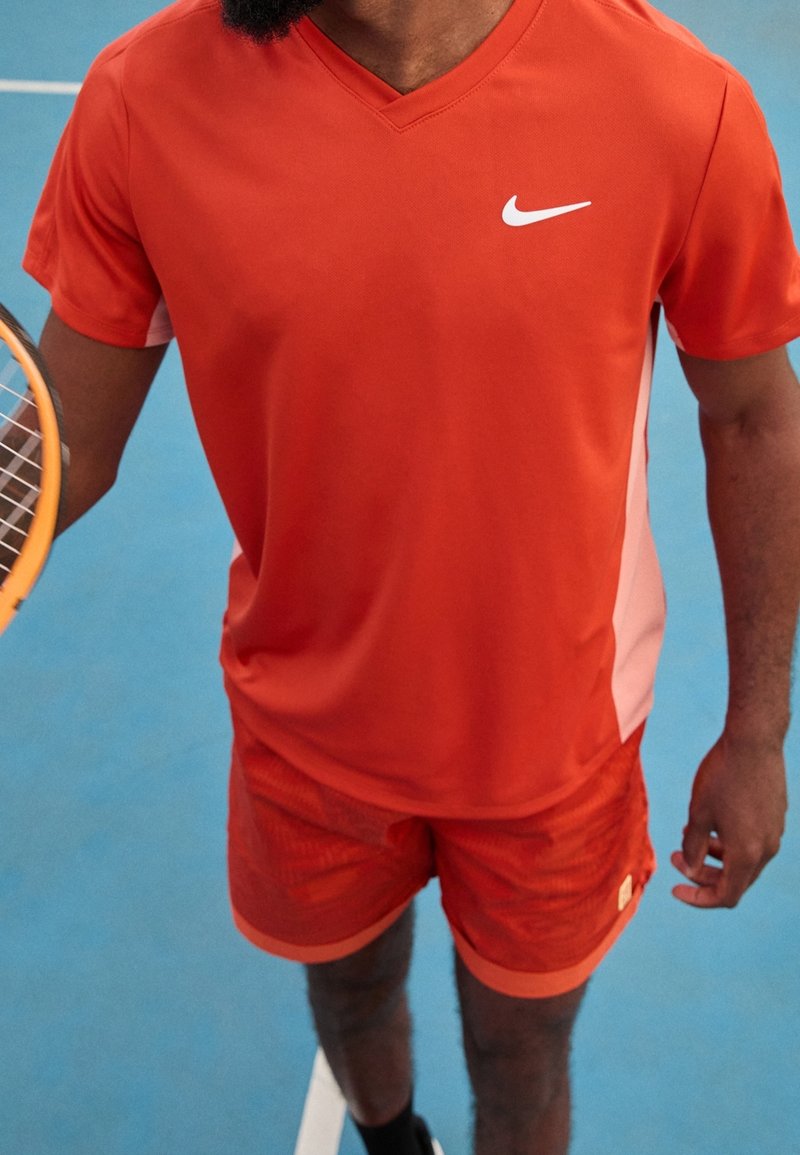 Sada výkonného sportovního oblečení v jasně oranžové barvě, zahrnující tričko s krátkým rukávem ve v-neck stylu s bílými detaily a odpovídající texturované šortky.