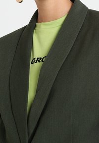 Blazer vert foncé avec un col châle, porté sur un t-shirt vert clair avec un texte noir. Texture de tissu lisse, design sur mesure.
