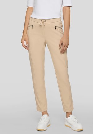 Beige, schmal zulaufende Hose mit vorderen Reißverschlusstaschen und Tunnelzugbund, getragen mit weißen Sneakern und einem weißen Kurzarmoberteil.
