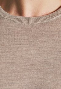 Gros plan sur le cou et le haut de la poitrine d'une personne portant un pull en maille beige à col rond, mettant en valeur la texture fine du tissu et le col côtelé.